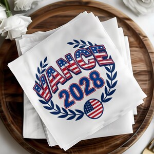 May include: White square napkins with the text "VANCE 2028" in red, white, and blue, surrounded by a blue laurel wreath and a small American flag emblem. The napkins are stacked on a wooden tray.