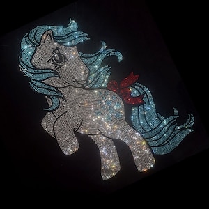 Puede incluir: Un diseño de poni con pedrería sobre un fondo negro. El poni es principalmente plateado con pelo azul claro y un lazo rojo. El diseño está hecho de muchas pedrerías pequeñas y brillantes.