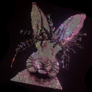 May include: A close-up of a sparkly pink and white rabbit design made of rhinestones. The rabbit's face is centered in the image, with its ears extending to the top and sides. The design is on a black background.