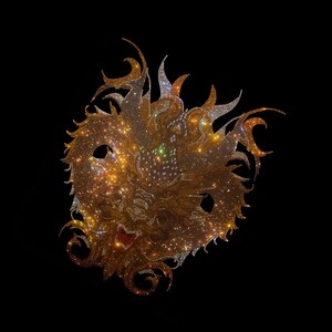 Puede incluir: Una máscara decorativa con un diseño similar a un león, cubierta de brillantes pedrería dorada y plateada. La máscara presenta agujeros para los ojos y detalles ornamentados en forma de llama en los bordes, sobre un fondo negro.