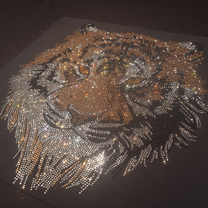 Op de afbeelding: Een close-up van een tijgergezicht gemaakt van strass. Het tijgergezicht is gemaakt van heldere en gouden strass, wat een sprankelend effect cre&euml;ert.