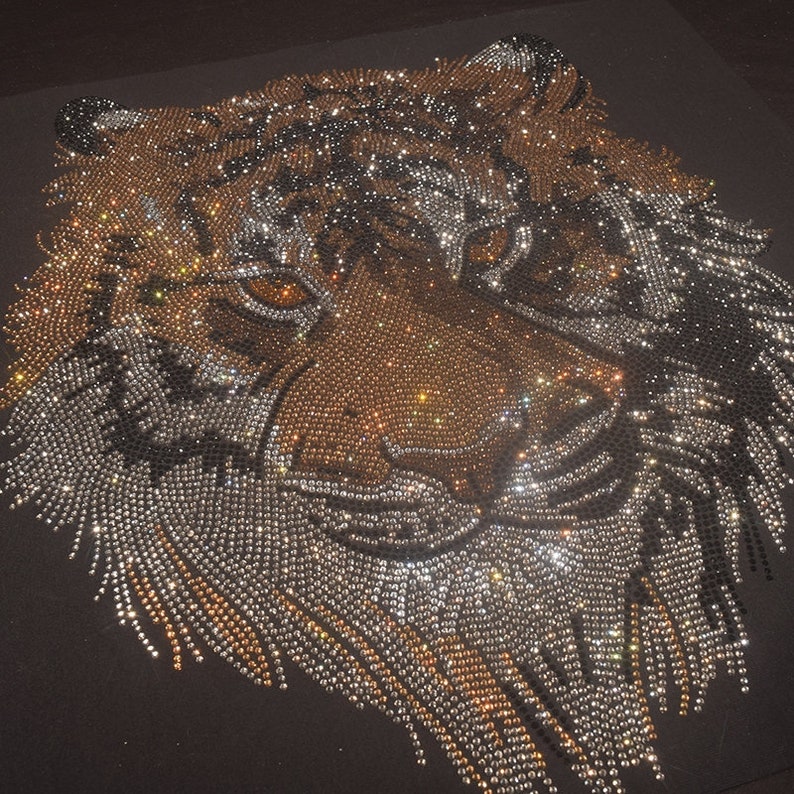 Op de afbeelding: Een close-up van een tijgergezicht gemaakt van strass. Het tijgergezicht is gemaakt van zilveren en gouden strass, wat een sprankelend effect cre&euml;ert.