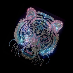Op de afbeelding: Een sprankelend tijgerkopontwerp, gemaakt met iriserende strass-steentjes. Het gezicht van de tijger heeft een mix van roze, blauwe en zilveren tinten, tegen een zwarte achtergrond. Het ontwerp bevat gedetailleerde gelaatstrekken en decoratieve strass-accenten.