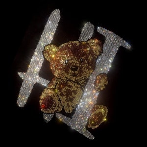 May include: A brown teddy bear with a sparkly gold design is surrounded by a sparkling silver 'H' design. The design is made of rhinestones.