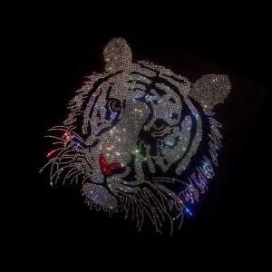 Peut inclure: Un visage de tigre blanc fait de strass sur un fond noir. Les yeux du tigre sont soulignés de noir et le nez est rouge.
