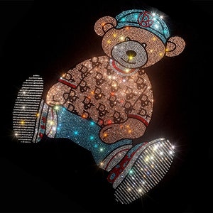 Puede incluir: Un oso de peluche cubierto de pedrería que lleva una gorra de béisbol azul y blanca, una camisa de patrón azul y negro y unos vaqueros azules. El oso está de pie sobre un fondo negro.