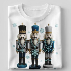 Puede incluir: Camiseta blanca con un diseño de tres cascanueces decorativos con uniformes azules, grises y negros. Cada cascanueces tiene un bigote blanco y detalles de copos de nieve. El diseño es festivo y de temporada.