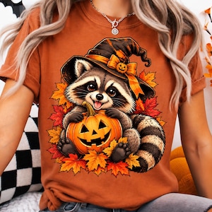 Peut inclure: Un t-shirt orange brûlé avec une illustration d'un raton laveur portant un chapeau de sorcière noir orné d'un nœud orange et d'une citrouille. Le raton laveur tient une citrouille d'Halloween sculptée entourée de feuilles d'automne rouges.