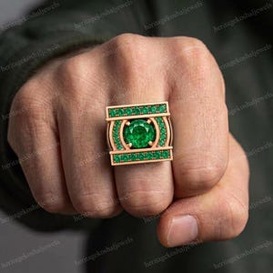 May include: A rose gold ring featuring a large, round, brilliant-cut green centre stone surrounded by a halo of smaller green stones. The rectangular setting includes two horizontal rows of green stones above and below the centre.