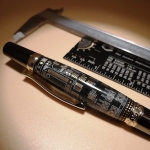 Puede incluir: Un bolígrafo negro con un diseño de placa de circuito, detalles plateados y un clip. Una regla negra con medidas en pulgadas y un calibrador plateado están en el fondo. El bolígrafo tiene el texto "CN" y "R122".