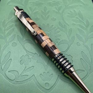 Könnte beinhalten: Ein Holzstift mit einem Schachbrettmuster und goldenen Akzenten. Der Stift hat ein Design aus schwarzem und braunem Holz und eine goldene Spitze.