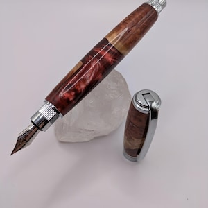 May include: A handcrafted fountain pen with a wooden barrel featuring a mix of reddish-brown and light brown hues. The pen has a silver-colored nib and metal accents. The cap is detached, revealing the pen's design.