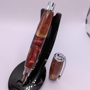 May include: A handcrafted fountain pen with a wooden barrel featuring a mix of reddish-brown and light brown tones. The pen has a silver-colored nib and accents, and is displayed on a black stand. The cap is detached and placed next to the pen.