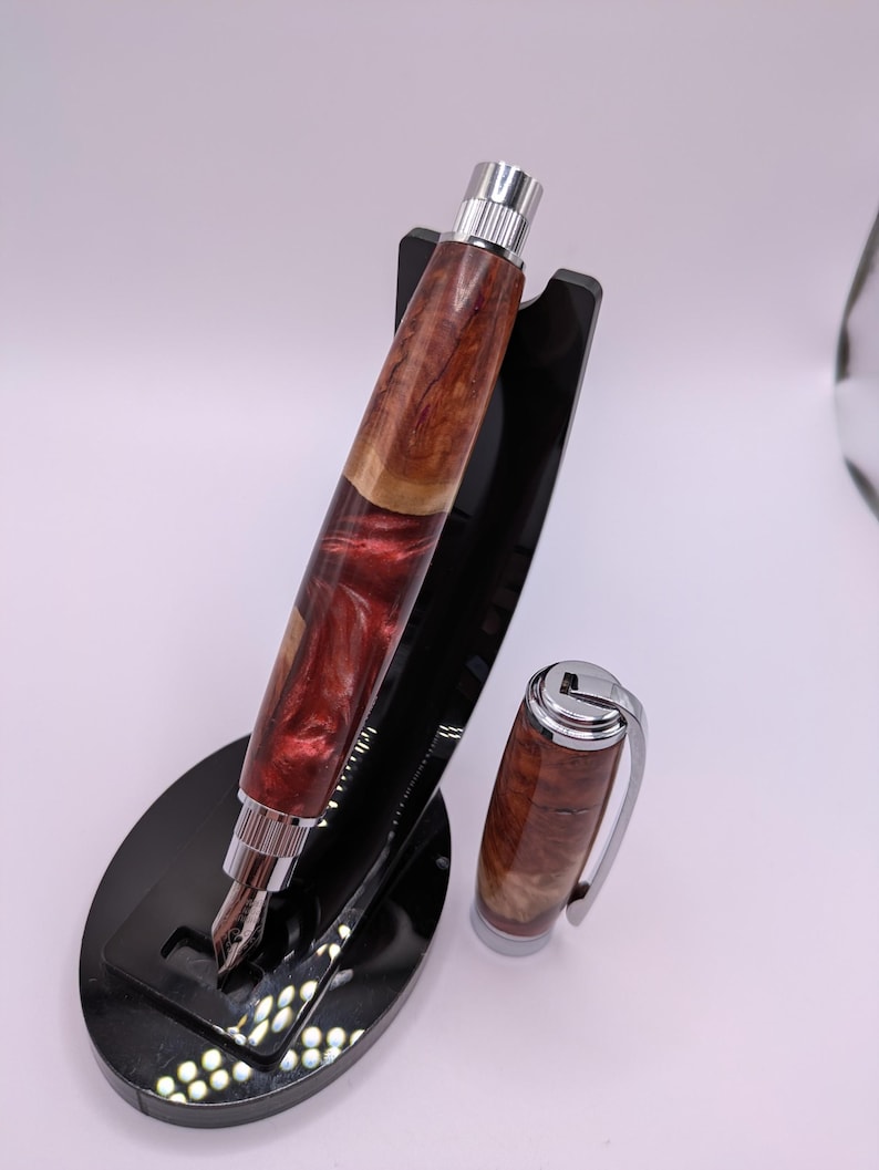 May include: A handcrafted fountain pen with a wooden barrel featuring a mix of reddish-brown and light brown tones. The pen has a silver-colored nib and accents, and is displayed on a black stand. The cap is detached and placed next to the pen.