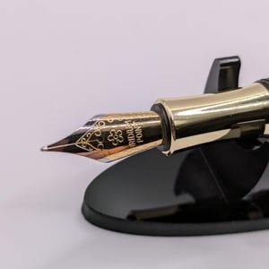 May include: Close-up of a gold-colored fountain pen nib with intricate floral designs and the text "IRIDIUM POINT". The pen is set on a black pen stand, showcasing its elegant design. The nib has a fine point.