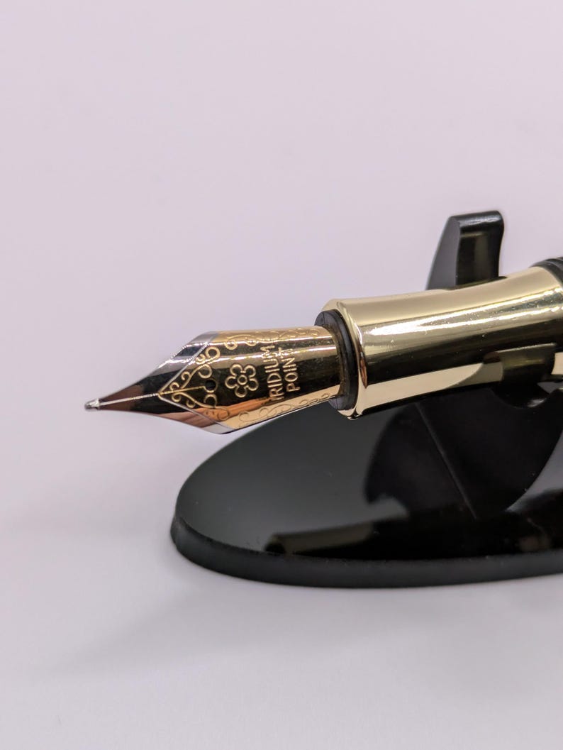 May include: Close-up of a gold-colored fountain pen nib with intricate floral designs and the text "IRIDIUM POINT". The pen is set on a black pen stand, showcasing its elegant design. The nib has a fine point.