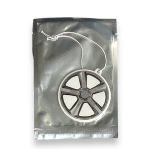 May include: A white car air freshener shaped like a wheel with a black rim and five spokes. The air freshener is packaged in a silver plastic bag with a white string.