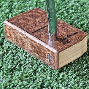 Könnte beinhalten: Ein hölzerner Golfschläger mit einem grünen Schaft und einem silbernen Kopf. Der Schläger ist mit den Worten "Bladewood #5" und einem kleinen Baumlogo graviert. Der Schläger liegt auf einer grünen künstlichen Grasfläche.