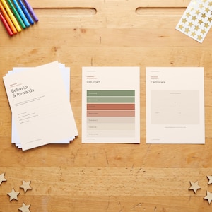 Op de afbeelding: Een set bedrukte vellen papier met een gedrags- en beloningsformulier, een clipkaart met gekleurde horizontale balken en een certificaatsjabloon, gerangschikt op een houten oppervlak met kleurrijke stiften en gouden sterstickers.