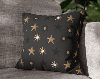 May include: A decorative throw pillow with a black background and a repeating pattern of gold, white, and dark gray stars and sunbursts. The pillow is square and rests on a gray sofa, adding a touch of celestial charm to home decor.
