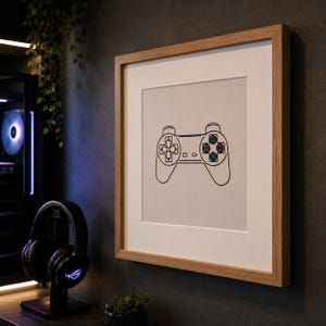 Op de afbeelding: Een vierkant ingelijst kunstwerk aan de muur met een minimalistische lijntekening van een videogamecontroller op een lichtgrijze achtergrond. Het kunstwerk zit in een lichthouten lijst met een witte passe-partout voor een strakke uitstraling.