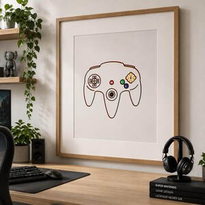 Puede incluir: Una lámina de arte mural enmarcada que presenta un dibujo lineal minimalista de un mando de videojuegos. El contorno del mando es negro con detalles de botones en rojo, amarillo, verde y azul. La lámina tiene un marco de madera clara.