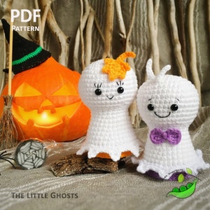 May include: Two crocheted ghost amigurumi toys with smiling faces. The ghost on the left has an orange leaf on its head. The ghost on the right has a purple bow tie. The ghosts are sitting in front of a lit jack-o-lantern and a small spider web.