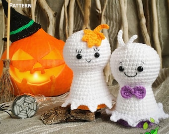 May include: Two crocheted ghost amigurumi toys with smiling faces. The ghost on the left has an orange leaf on its head. The ghost on the right has a purple bow tie. The ghosts are sitting in front of a lit jack-o-lantern and a small spider web.