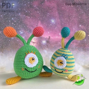 Puede incluir: Dos monstruos de crochet con forma de huevo con un ojo grande cada uno. Un monstruo es verde con antenas naranjas y rosas, el otro es amarillo y rayado con antenas azules y amarillas. Ambos monstruos tienen un ojo blanco con una pupila amarilla y una boca sonriente.