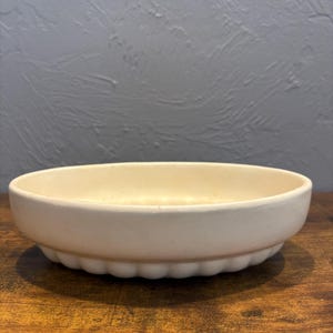 May include: A cream-colored, ceramic serving dish with a scalloped edge. The shallow, oval-shaped dish sits on a wooden surface against a gray wall. The dish is empty and appears to be new.