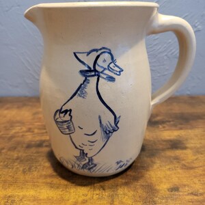 May include: A white ceramic pitcher with a blue hand-painted design of a duck carrying a bucket. The duck is wearing a bonnet and has a blue collar.