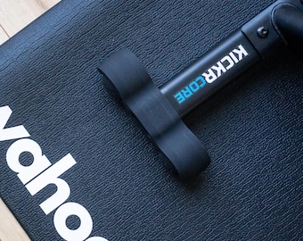 May include: Black exercise mat with the white word "Wahoo" printed on it. A black exercise bike component is on the mat, with the words "KICKR CORE" in white and blue. The mat has a textured surface.