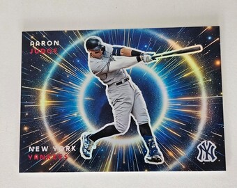 May include: Baseball card featuring Aaron Judge of the New York Yankees in a batting pose. The card has a vibrant background with a radial design in shades of blue, yellow, and orange. The Yankees logo is visible, along with Judge's name.