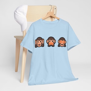 Peut inclure: T-shirt bleu clair avec un motif de trois singes, l'un se couvrant les oreilles, l'autre se couvrant la bouche et le dernier se couvrant les yeux. Les singes sont jaunes avec des détails orange.