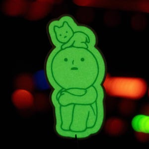 May include: A green glow-in-the-dark sticker of a small person with a cat sitting on its head. The person is sitting with its arms crossed and has a sad expression.
