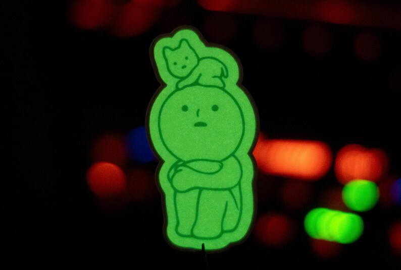 May include: A green glow-in-the-dark sticker of a small person with a cat sitting on its head. The person is sitting with its arms crossed and has a sad expression.