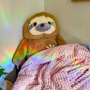 May include: A brown sloth plush toy is lying under a pink and white striped blanket. The blanket is made of a soft, fluffy material. The sloth has a gray face with black eyes and a black nose.