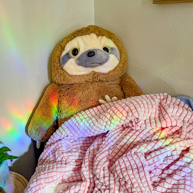May include: A brown sloth plush toy is lying under a pink and white striped blanket. The blanket is made of a soft, fluffy material. The sloth has a gray face with black eyes and a black nose.