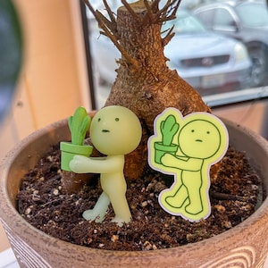 May include: Two green glow-in-the-dark figurines holding small potted plants. The figurines are standing in a brown pot filled with soil. The pot has a decorative pattern on the side.