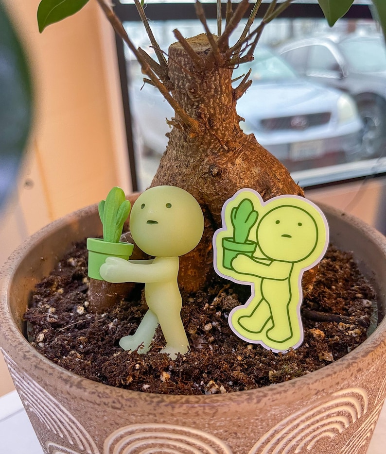 May include: Two green glow-in-the-dark figurines holding small potted plants. The figurines are standing in a brown pot filled with soil. The pot has a decorative pattern on the side.