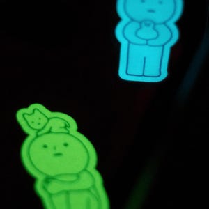 May include: Two glow-in-the-dark stickers featuring simple cartoon characters. The first sticker is blue and depicts a person holding a small animal. The second sticker is green and depicts a person with a small animal on their head.