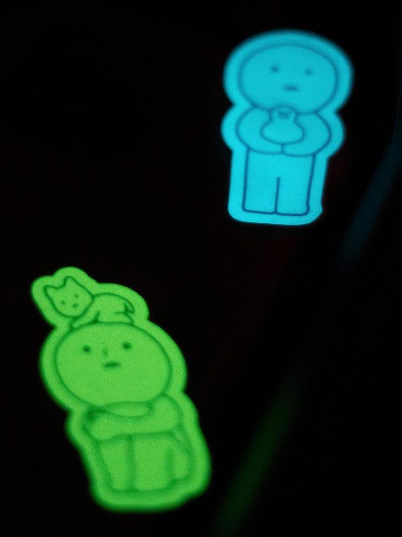 May include: Two glow-in-the-dark stickers featuring simple cartoon characters. The first sticker is blue and depicts a person holding a small animal. The second sticker is green and depicts a person with a small animal on their head.