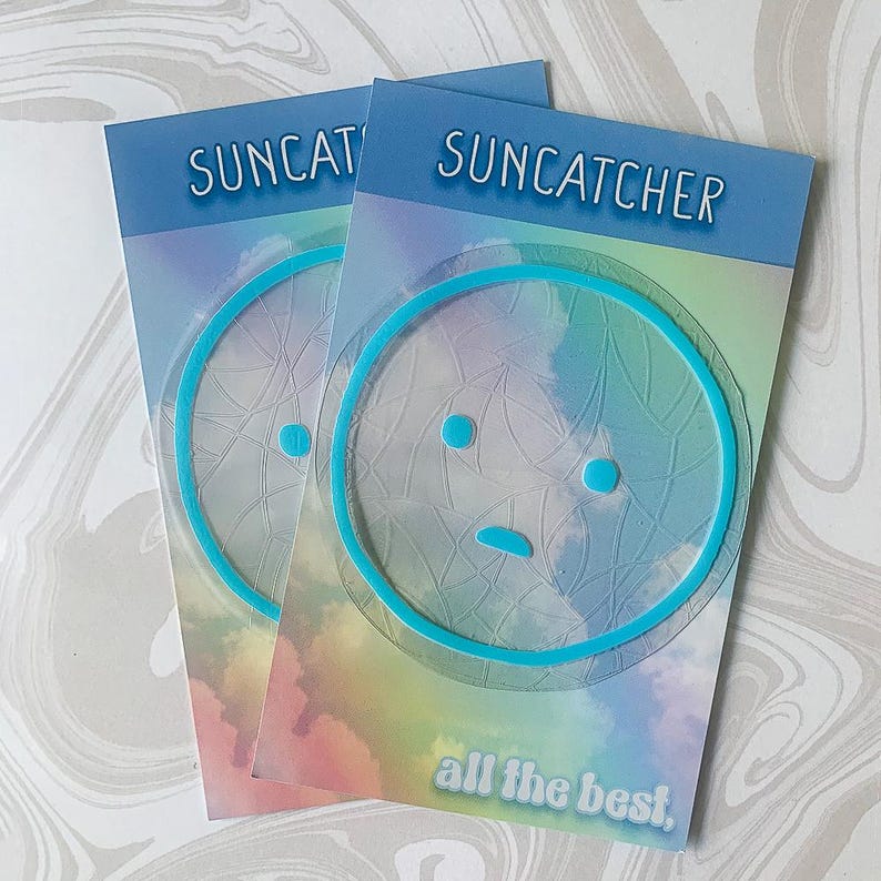 May include: Two pastel blue and purple cards with the text "SUNCATCHER" and "all the best."  Each card features a blue outlined sticker of a cartoon face with a sad expression.