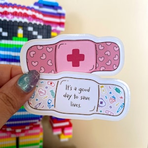 May include: Two pink and white band-aid stickers. The top sticker has a red plus sign and pink hearts. The bottom sticker has the text "It's a good day to save lives." and is filled with small medical icons.