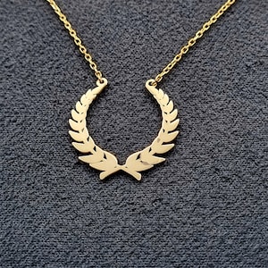 May include: A gold necklace with a laurel wreath pendant. The necklace is on a dark gray background.