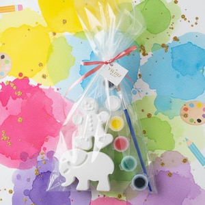 May include: A clear cellophane bag tied with a pink ribbon contains a paint-your-own kit. The kit includes a white gingerbread man and elephant figurine, paint pots, and a paintbrush. The background features colorful watercolor paint splatters and gold glitter.