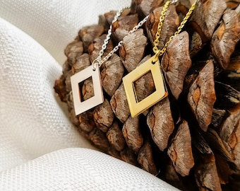 May include: Two delicate necklaces with square pendant charms. One necklace has a silver charm and the other has a gold charm. The necklaces are displayed on a brown pine cone.