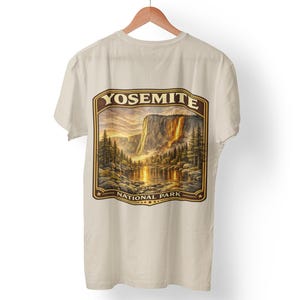 May include: Cream-colored t-shirt featuring a scenic illustration of Yosemite National Park. The design includes a waterfall, lake, and trees under a sunset sky, with the words "YOSEMITE NATIONAL PARK" in a framed border.