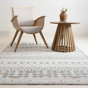 Può includere: Una sedia in legno chiaro con un cuscino bianco si trova su un tappeto a motivi grigi e bianchi con un tavolino in legno chiaro con un vaso intrecciato e fiori bianchi in cima.