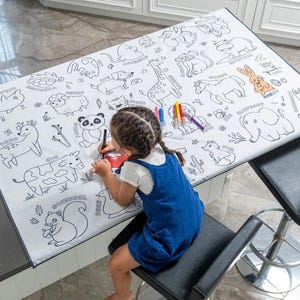 May include: A white tablecloth with black outlines of various animals, including a deer, cow, squirrel, duck, panda, fox, elephant, giraffe, and more. A child is coloring the tablecloth with markers.